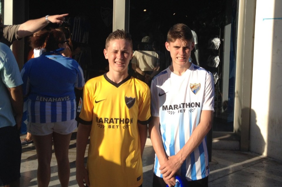 Two boys in soccer jerseys stand outside a shop.