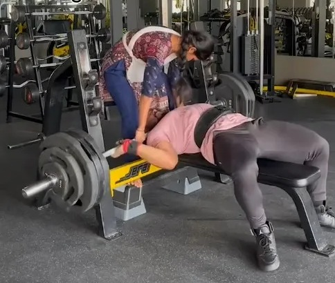 A woman bench pressing 165kg in a gym, assisted by another woman.