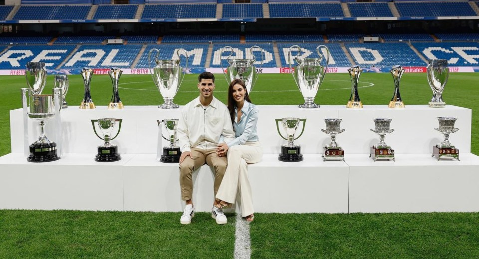 Marco Ascensio and Sandra Garal sitting with trophies in front of a Real Madrid stadium.