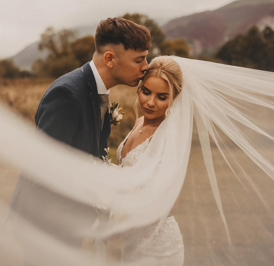 Groom kissing bride on forehead, veil flowing in wind.