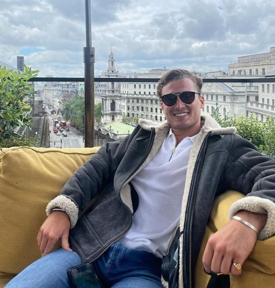 Man in a shearling jacket sitting on a rooftop overlooking a city.