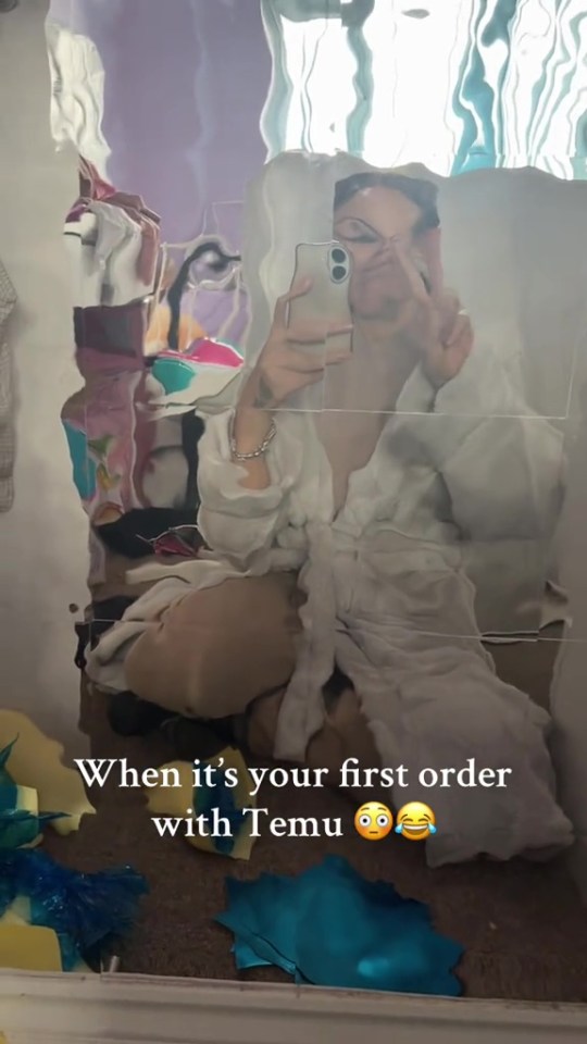 Woman in robe taking selfie in distorted mirror, surrounded by scattered items from a Temu order.