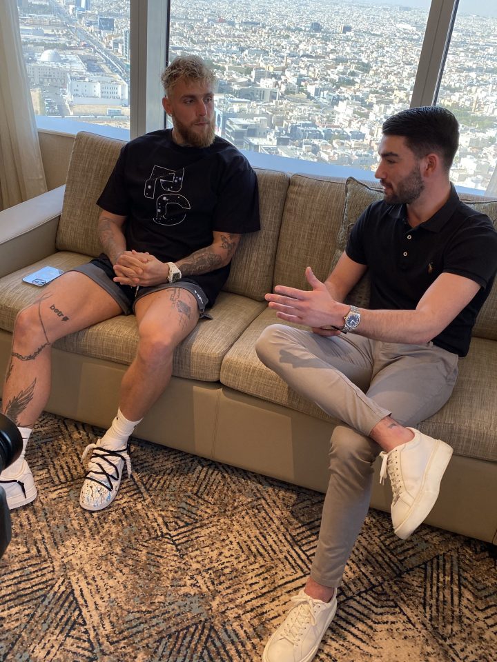 Two men sitting on a couch, talking.  A city is visible through a large window behind them.