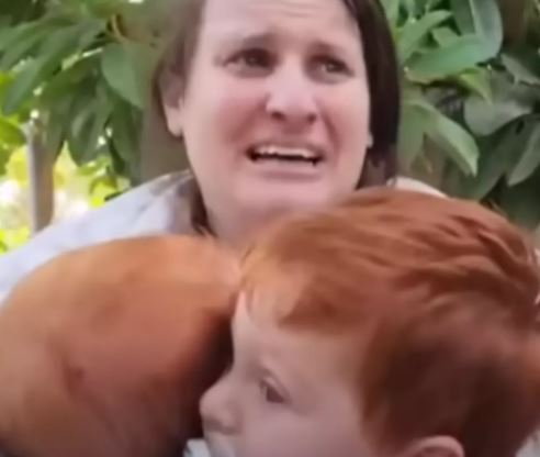 A woman holding two young children, appearing upset.