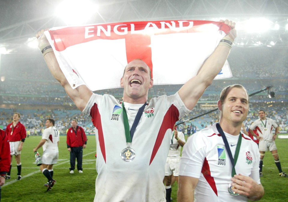 Two rugby players celebrate an England victory, holding medals and a flag.