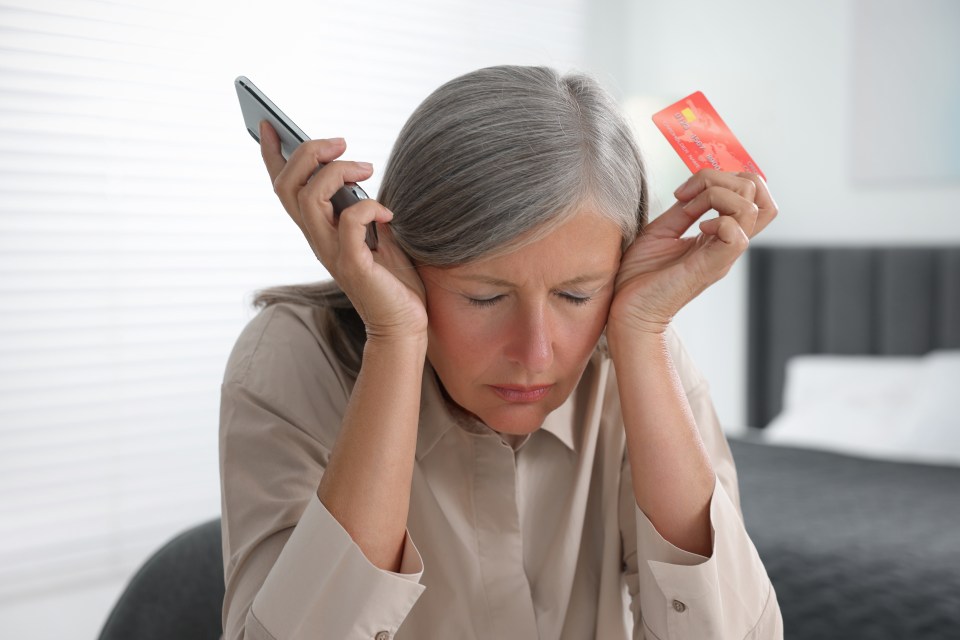 Distraught senior woman holding a smartphone and credit card, victim of online fraud.