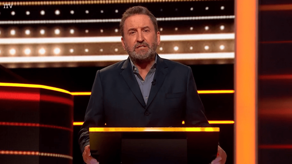 A man in a suit standing at a podium on a game show set.