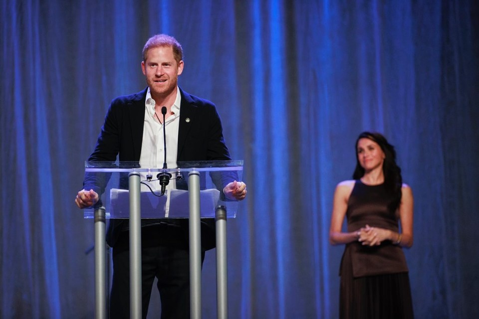 Prince Harry speaking at a podium with Meghan Markle standing behind him.