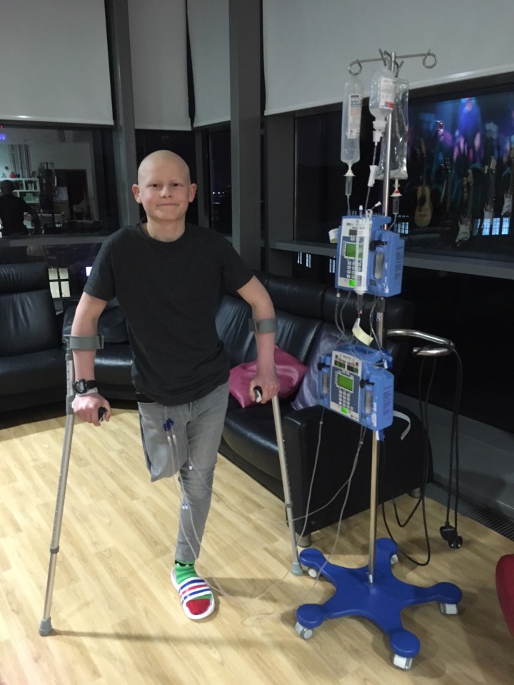 A boy with no hair uses crutches while connected to an IV pole.