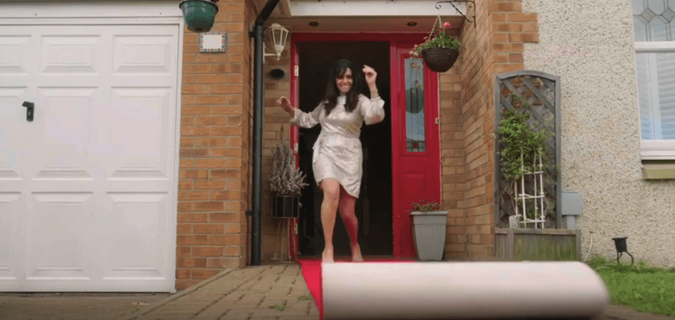 Woman in a silver dress joyfully unrolling a red carpet in front of her house.