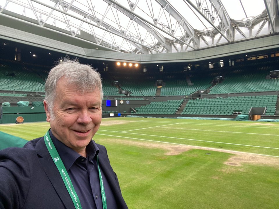 Man in a blazer smiles for a selfie in a nearly empty tennis stadium.