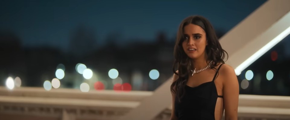 A young woman in a black dress stands on a bridge at night.