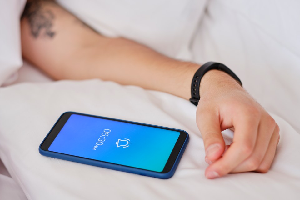 A smartphone alarm displaying 6:30 AM is on a bed next to a man's arm.