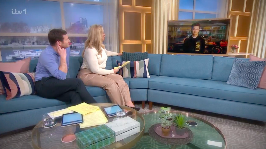 Screenshot of two people sitting on a sofa in a TV studio, watching a racing driver on a screen.