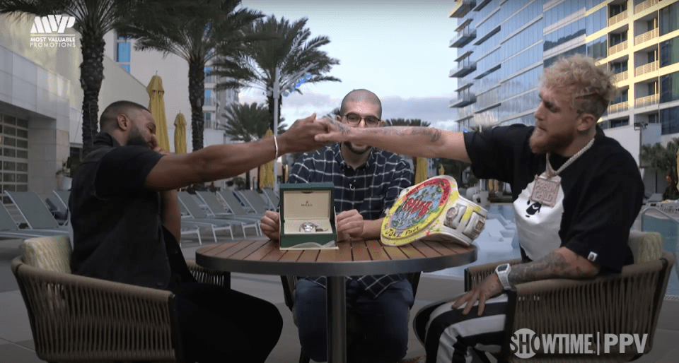 Three men sitting at a table outdoors, showcasing a watch and championship belt.