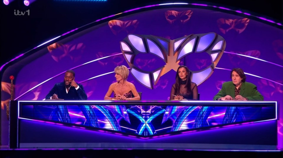 Screenshot of four judges sitting at a table in front of a large mask.