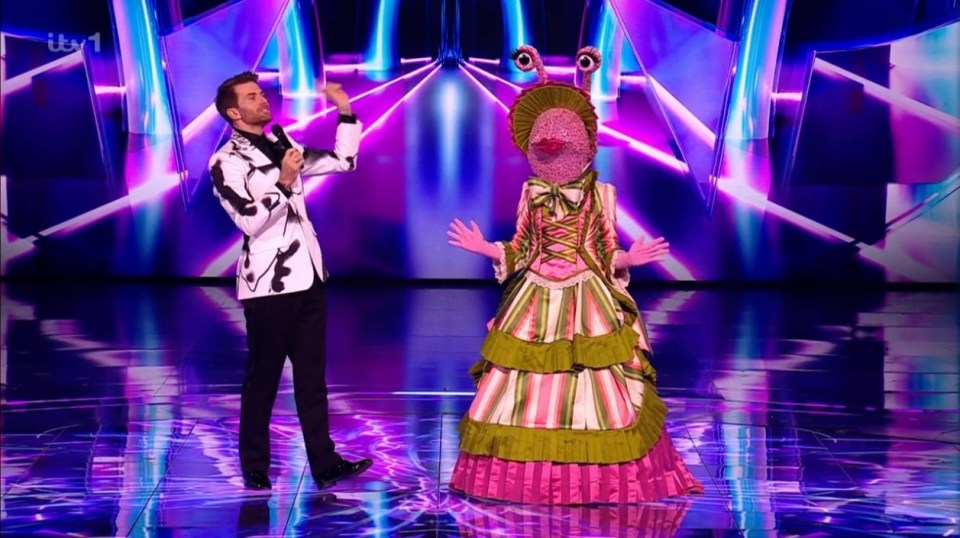 Screenshot of a masked singer contestant in a pink and green dress and a host on a stage.