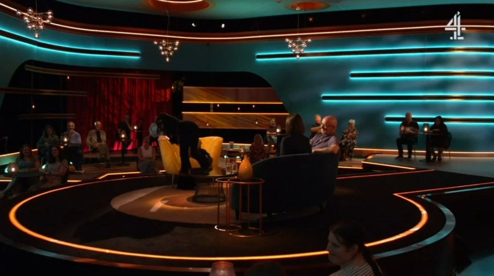 Screenshot of a television studio audience and guests seated in a curved, modern set.