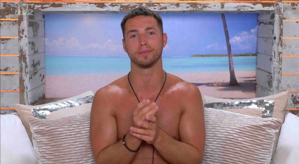 Shirtless man sitting on a bed, tropical beach scene in the background.