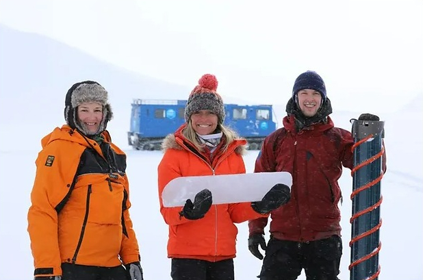 Three people in winter clothing hold a large piece of ice and a piece of equipment in a snowy landscape.