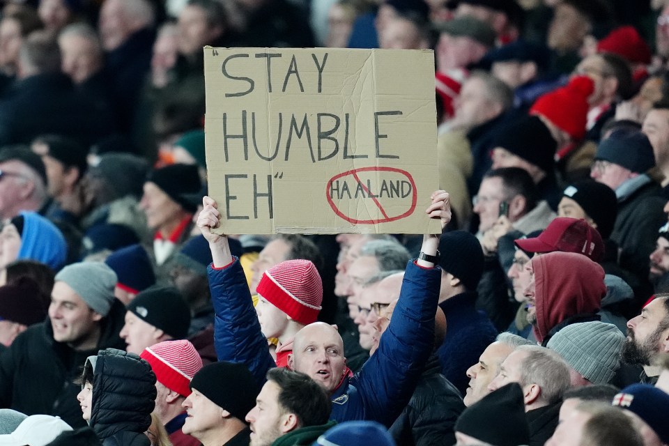 Arsenal fan holding a sign that says "Stay Humble Eh" with "Haaland" crossed out.