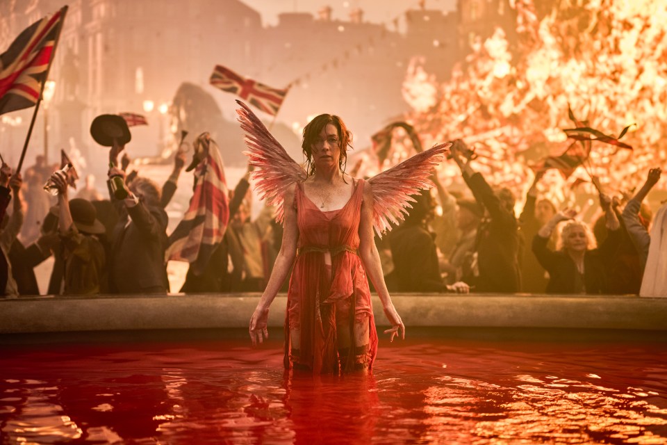 A woman with feathered wings stands in a pool of red water, surrounded by a crowd waving British flags.