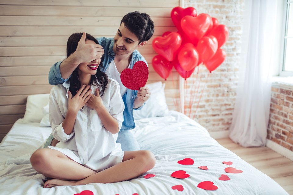 A young man surprises his girlfriend in bed on Valentine's Day.