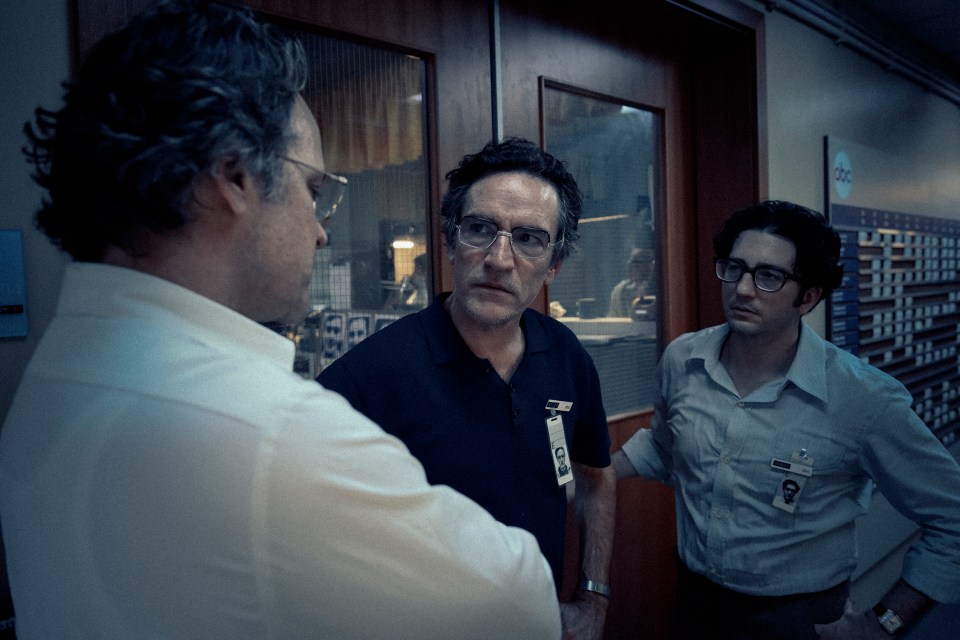 Film still of three men in a hallway, two in shirts and one in a polo shirt.