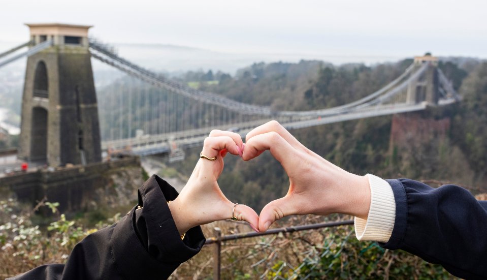 Two hands forming a heart shape in front of the Clifton Suspension Bridge.