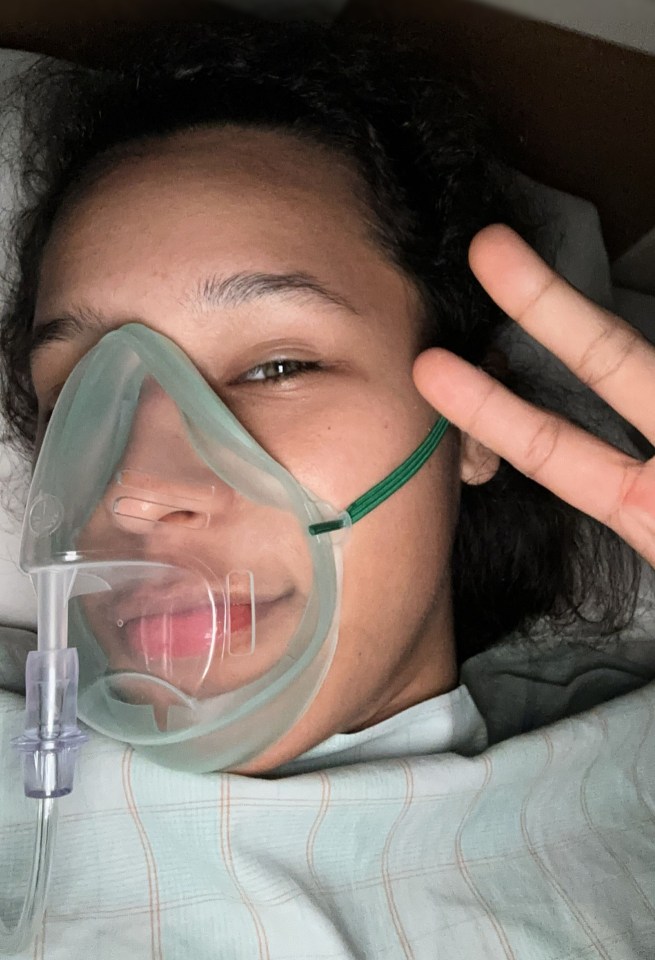 Woman in hospital bed wearing oxygen mask, giving peace sign.