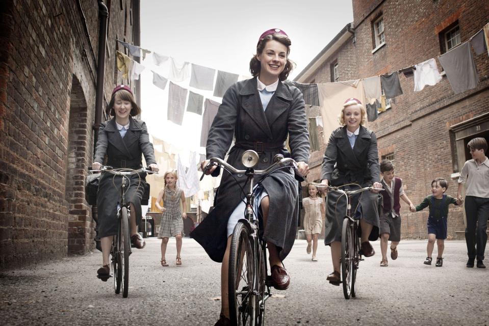 Three midwives on bicycles riding down a street, children running alongside.