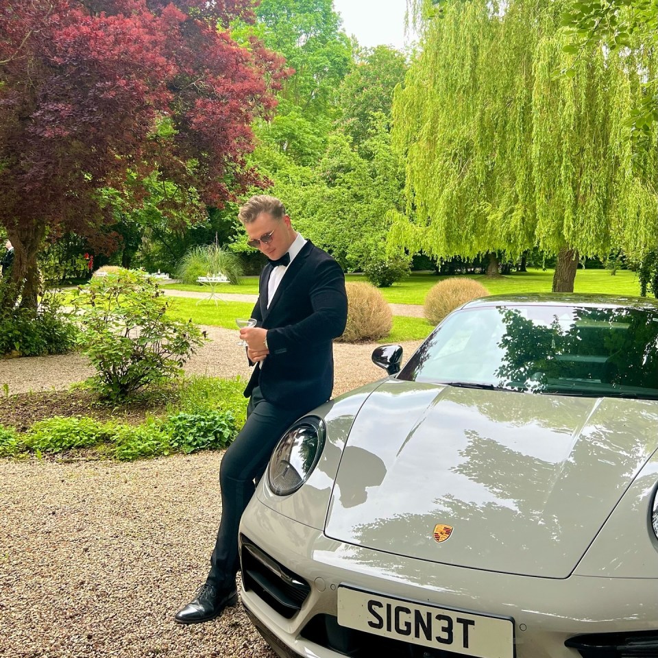 Man in tuxedo holding a glass stands next to a light gray Porsche.