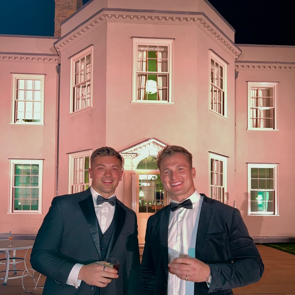 Two men in tuxedos stand outside a large house at night.