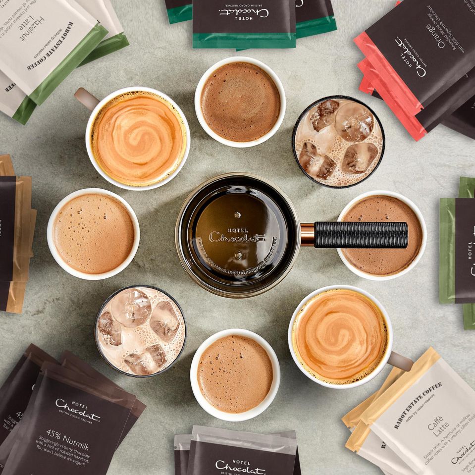 Hot chocolate in various mugs and glasses surrounding a copper-colored hot chocolate maker.