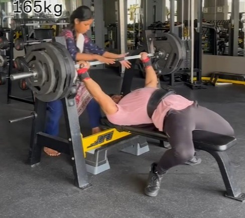 A woman spots a man bench pressing 165 kg in a gym.