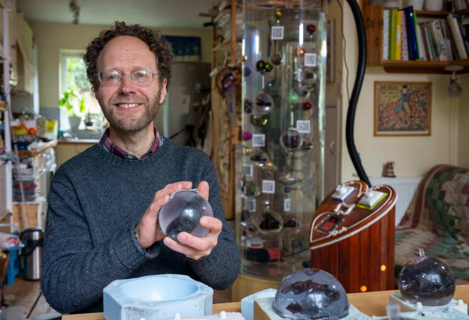 A man holding a glass sphere containing human feces, in front of a human poo aquarium.