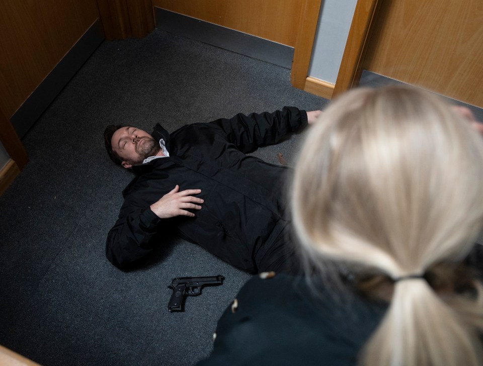 A man lies motionless on an office floor with a gun nearby; a woman looks down at him.