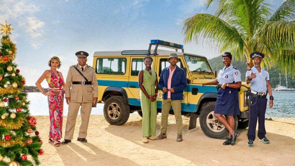 Cast photo from Death in Paradise Christmas special, featuring a police vehicle and a Christmas tree.