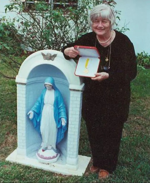 Woman holding a medal in front of a statue of the Virgin Mary.