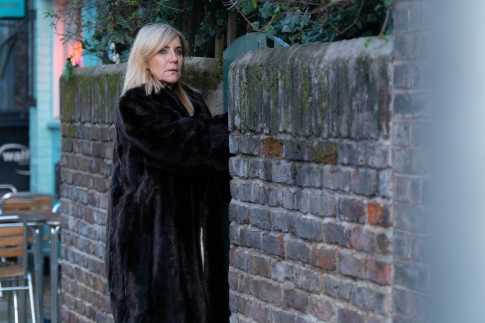 Michelle Collins as Cindy Beale in a dark fur coat, standing by a brick wall.