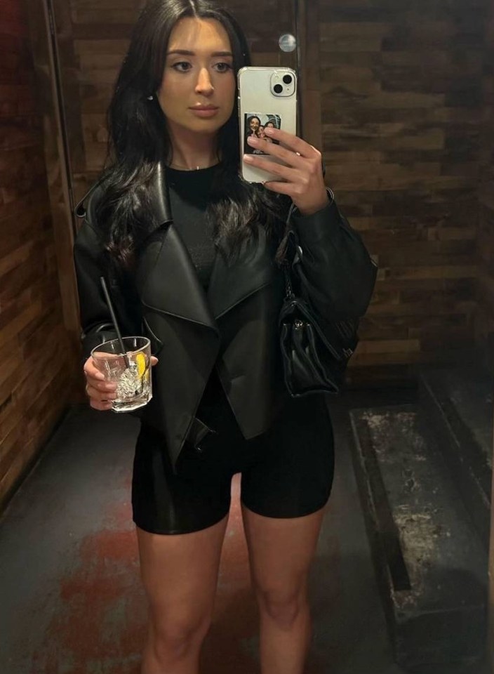 Woman in black biker jacket and shorts taking a selfie in a bar restroom.