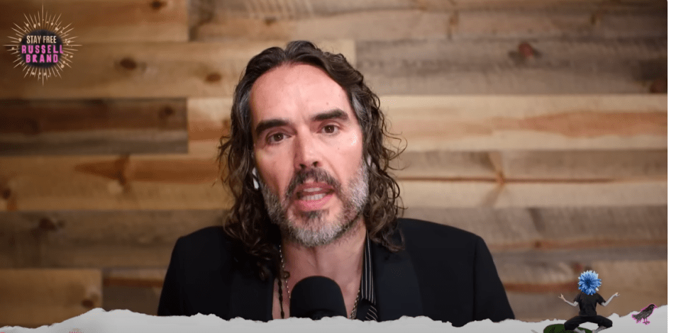 Russell Brand speaking in front of a wooden wall.