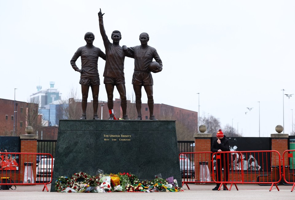 Statue of the United Trinity (Best, Law, Charlton) at Old Trafford with floral tributes.