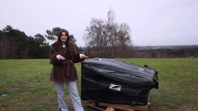 Woman standing in a field next to a large pallet of furniture wrapped in black plastic.