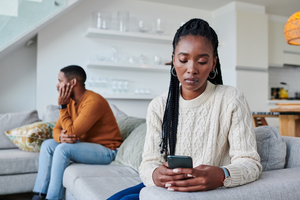 A woman sits on a couch looking at her phone while a man sits beside her looking upset.