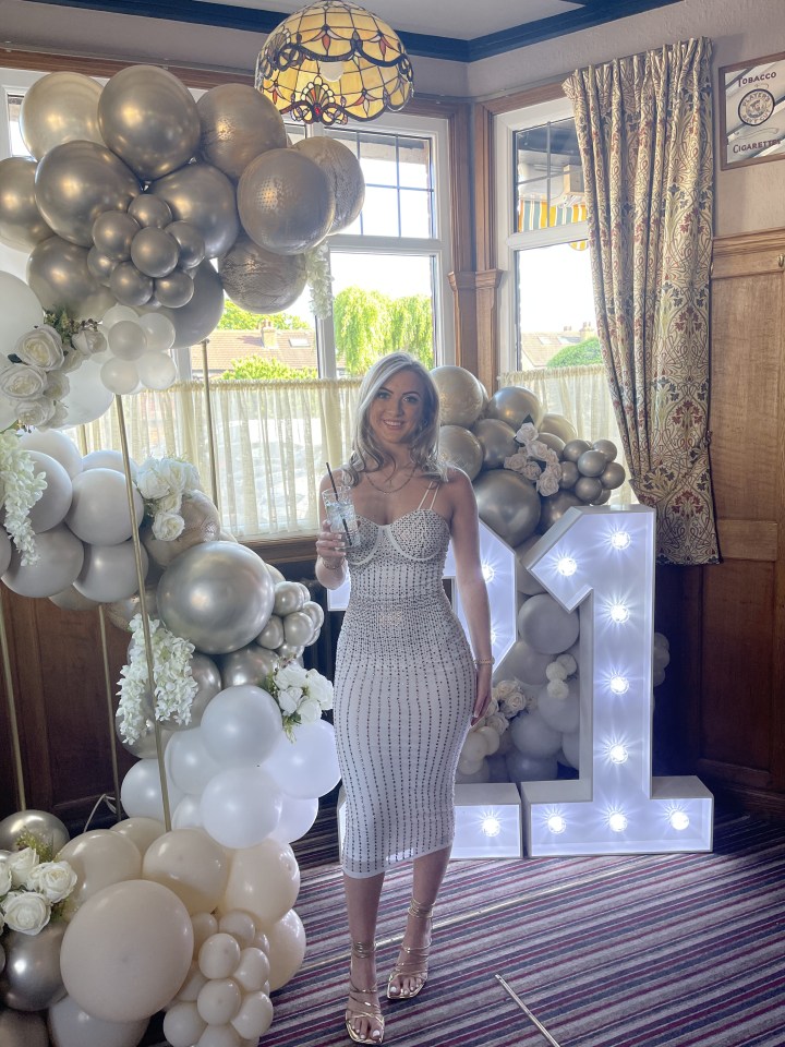 Woman in a white dress stands in front of a balloon arch and a lit number one.