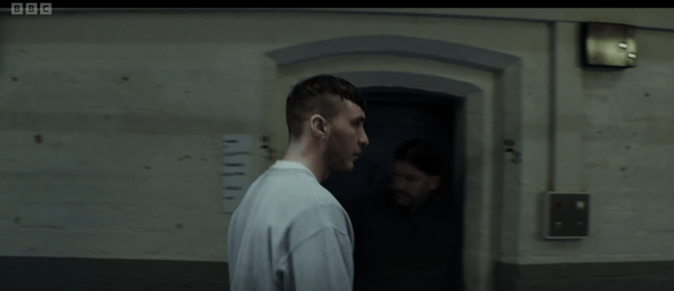 A man in a light grey prison uniform walks past a doorway, another man is visible in the background.