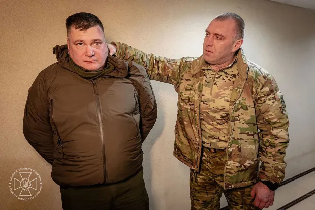 Photo of a man in a brown jacket being detained by a soldier.