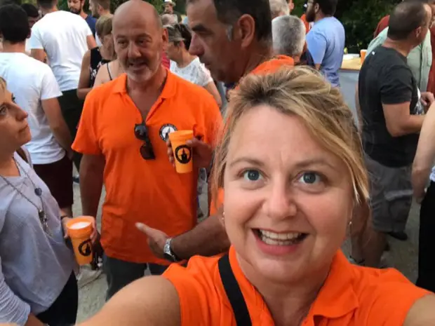 A woman in an orange shirt takes a selfie with people in the background.