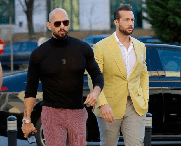 Two men walking, one in a black turtleneck and pink pants, the other in a yellow blazer and gray pants.